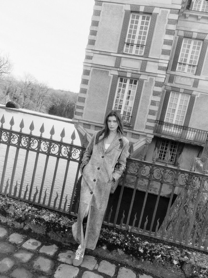 Black and white shot of a woman in a long coat standing by a railing with a building and trees in the background, fashion editorial.