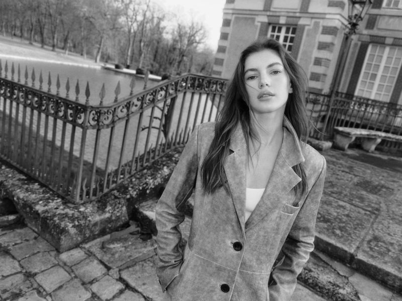 Black and white portrait of a woman wearing a jacket standing on a stone staircase, with a stone building in the background.