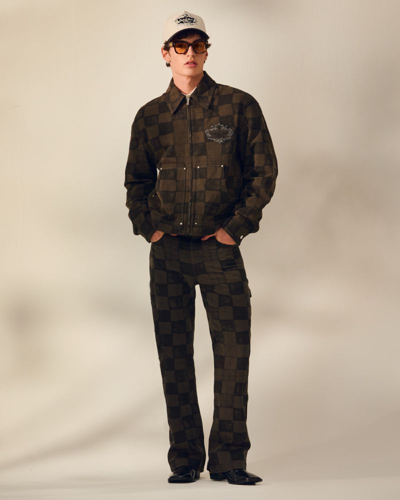 Full-length shot of a young adult man in a checkered jacket and pants with sunglasses and a baseball cap posing in a studio.