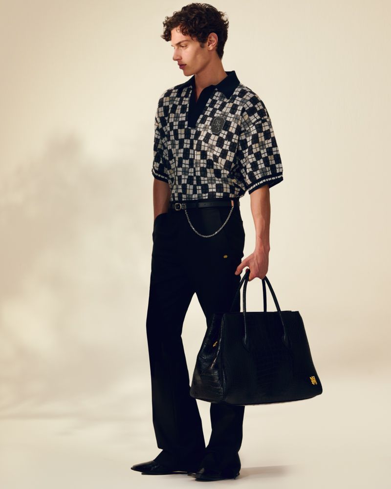 A stylish adult male stands indoors, dressed in black pants and a checkered shirt, holding a black leather tote bag in studio lighting.