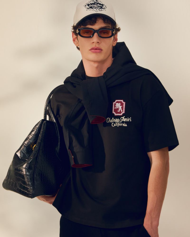 Man poses wearing a black t-shirt with a logo, a white baseball cap, and sunglasses, with a black handbag hanging from his shoulder.