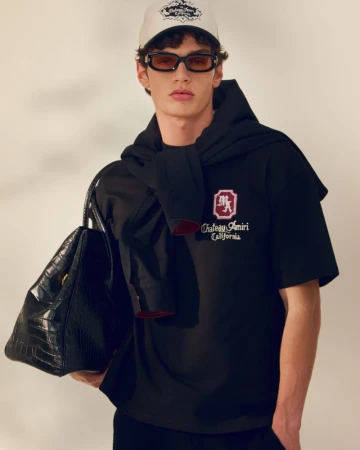 Man poses wearing a black t-shirt with a logo, a white baseball cap, and sunglasses, with a black handbag hanging from his shoulder.