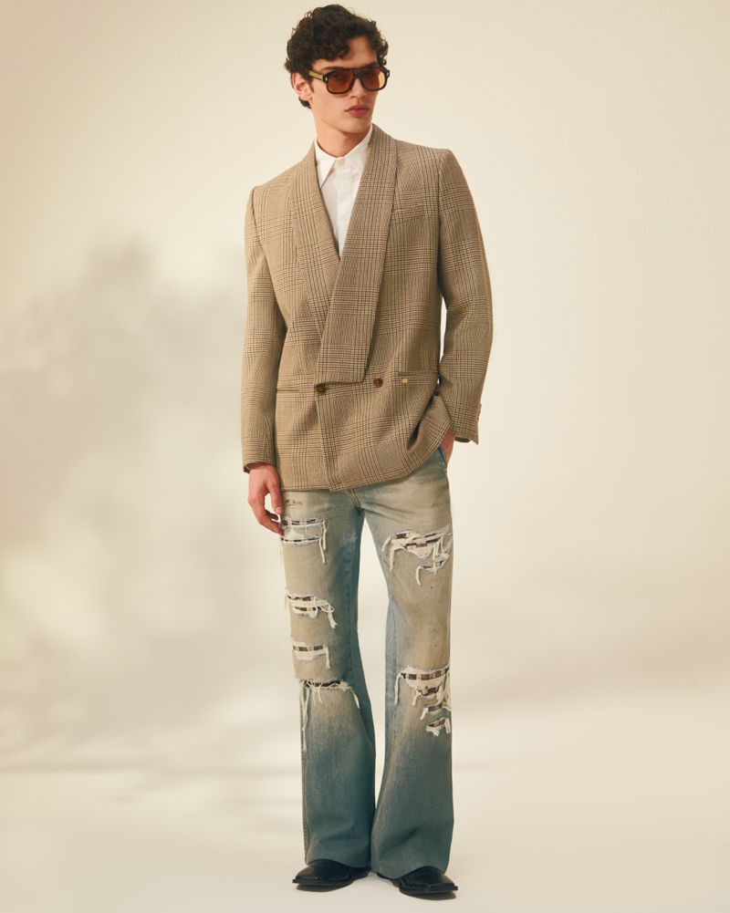 A man wearing a tan plaid blazer, white shirt, and heavily distressed light-wash flared jeans stands against a pale neutral background.