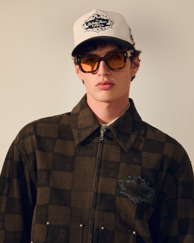 Male fashion model wearing a checkered jacket, cap, and sunglasses in a studio shot. Portrait-style image with neutral background.