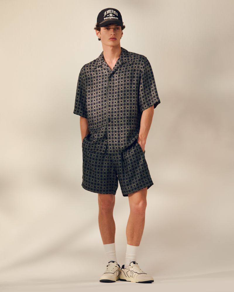 Man wearing a patterned shirt and shorts with white sneakers and a baseball cap, standing indoors against a neutral backdrop.