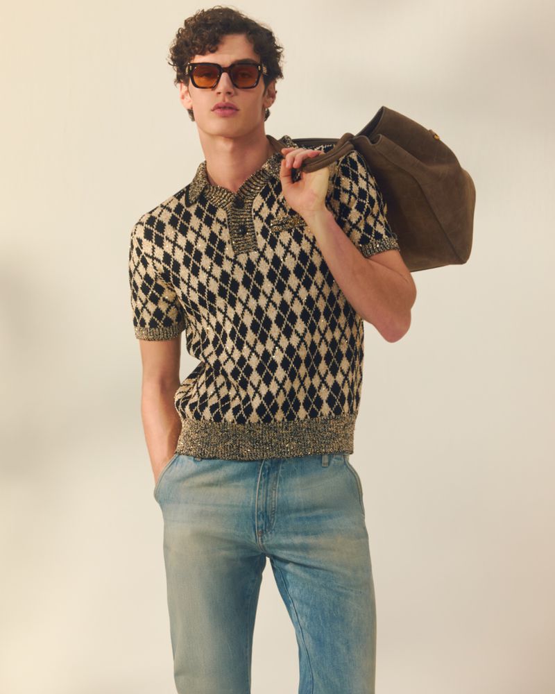 Model wearing dark sunglasses, patterned polo shirt, and light denim jeans while carrying a dark shoulder bag against a neutral background.