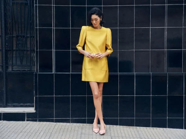 Portrait of a woman in a yellow dress and light pink heels posing against a black tile wall in an outdoor setting on a cloudy day.