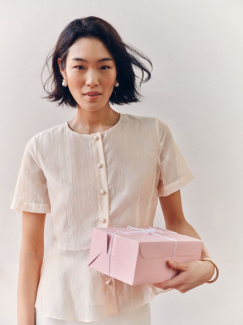 Young woman holding pink gift box with white ribbon. She wears a cream-colored blouse and stands against a neutral background in soft light.