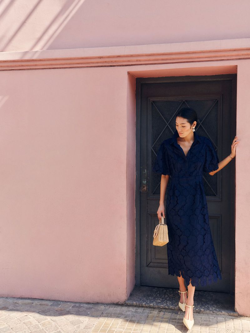 Elegant woman in blue floral dress and white heels poses beside a pink wall and grey door holding a straw purse in daylight.