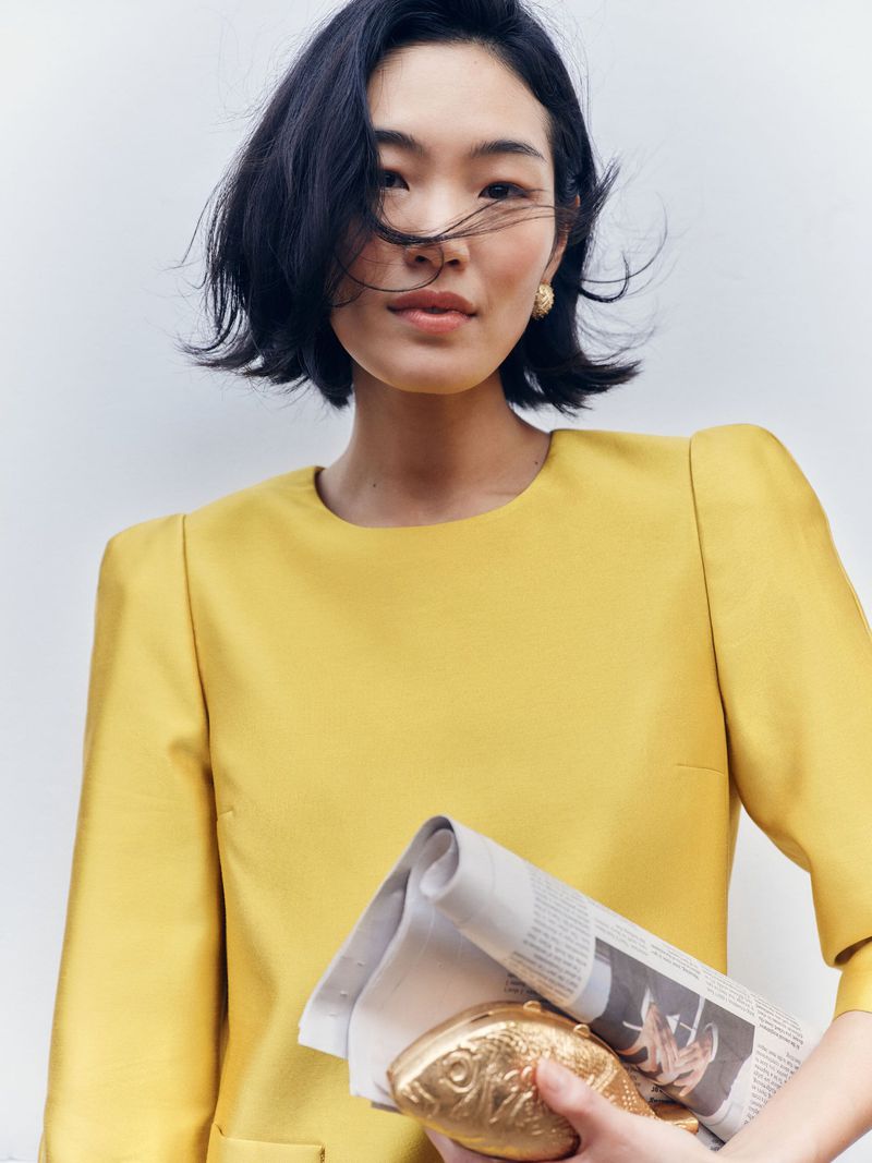 Asian woman in yellow dress holds newspaper and gold fish purse. Portrait shot with shallow depth of field, neutral background.