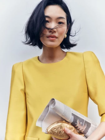 Asian woman with dark bob hair wearing a yellow dress holding a gold fish clutch and rolled newspaper against a white background