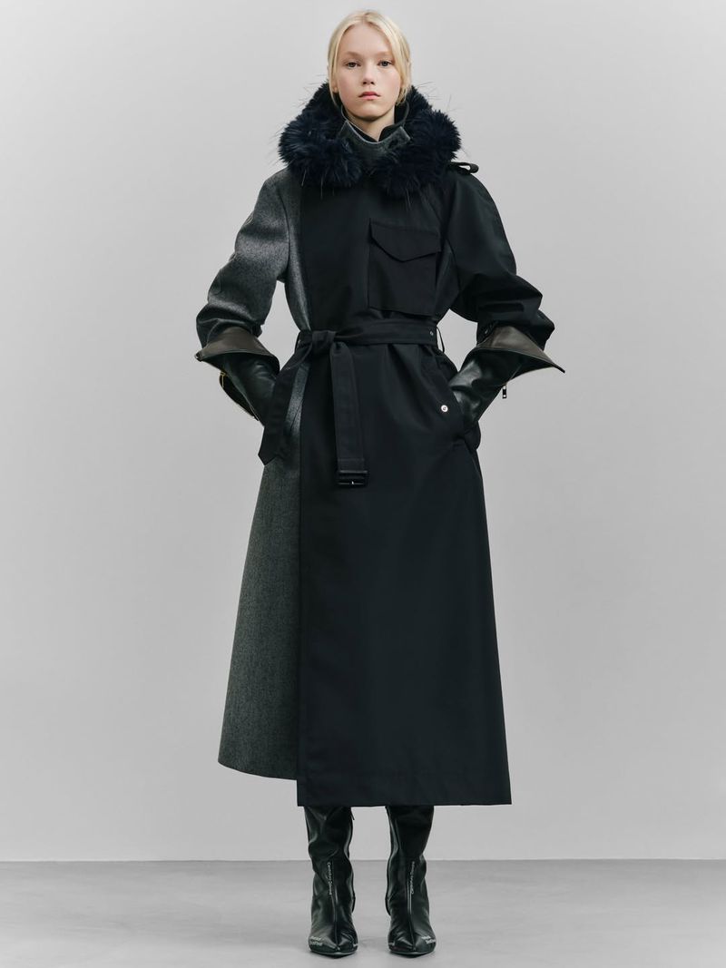 A woman stands in a studio wearing a long, belted mixed-fabric coat with a fur collar and black leather boots against a grey background.