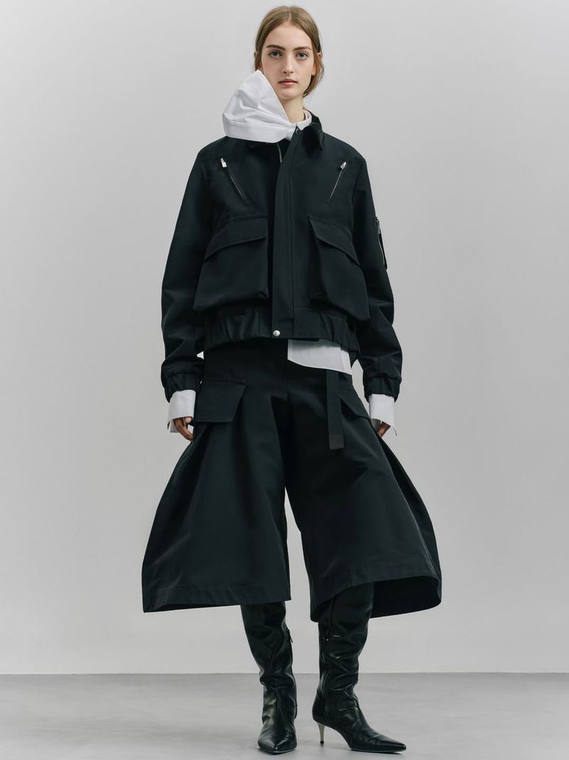 Fashionable adult female poses in black jacket and wide-leg pants with boots against a neutral backdrop in a full-length studio shot.