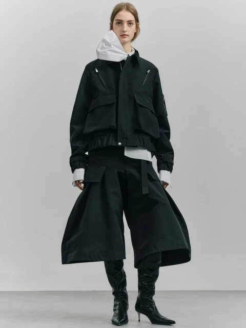 Fashionable adult female poses in black jacket and wide-leg pants with boots against a neutral backdrop in a full-length studio shot.