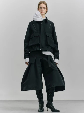 Fashionable adult female poses in black jacket and wide-leg pants with boots against a neutral backdrop in a full-length studio shot.