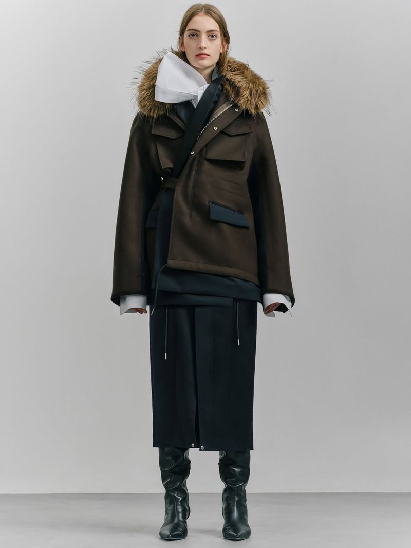 A woman in a fur-trimmed parka, layered top, and black skirt and boots poses in a studio. The outfit is styled with a mix of formal and casual elements.