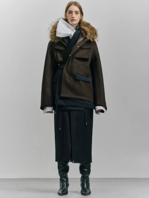 A woman in a fur-trimmed parka, layered top, and black skirt and boots poses in a studio. The outfit is styled with a mix of formal and casual elements.