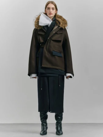 A woman in a fur-trimmed parka, layered top, and black skirt and boots poses in a studio. The outfit is styled with a mix of formal and casual elements.