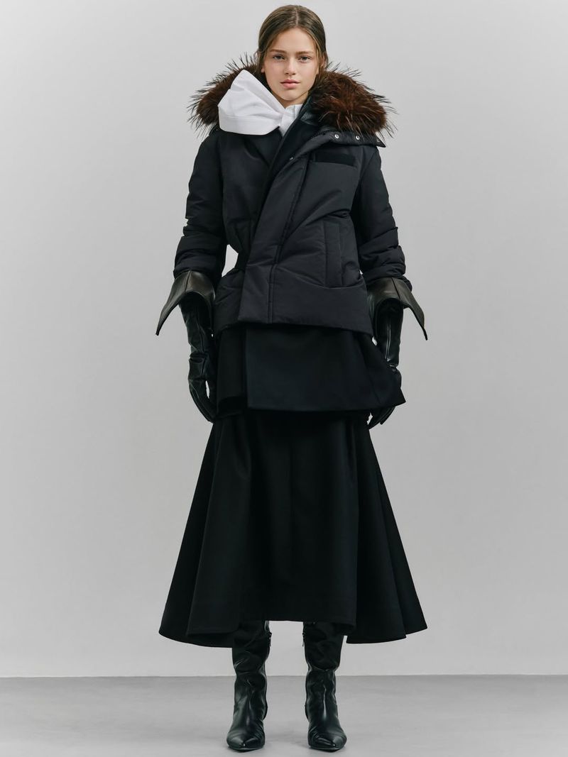 A woman stands in a studio wearing a black down jacket with fur trim, a white hooded sweatshirt, a black skirt, and black leather boots.