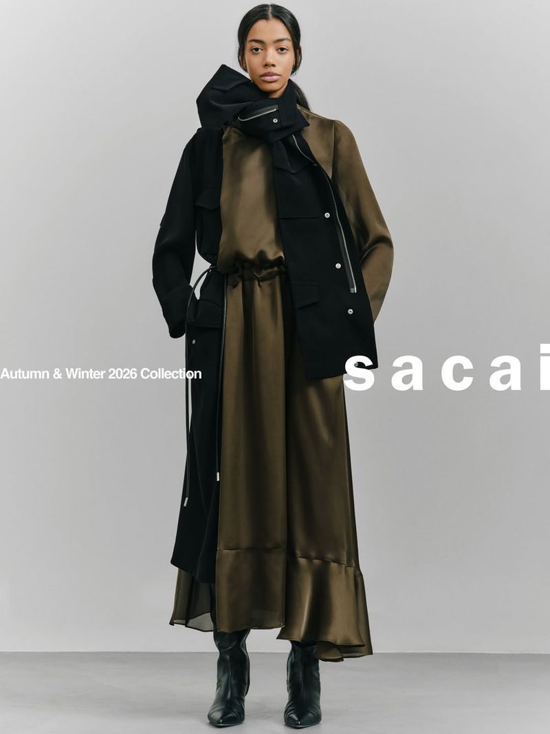A woman stands in a fashion-forward pose wearing a black jacket and olive green dress in a studio setting with soft, neutral lighting.