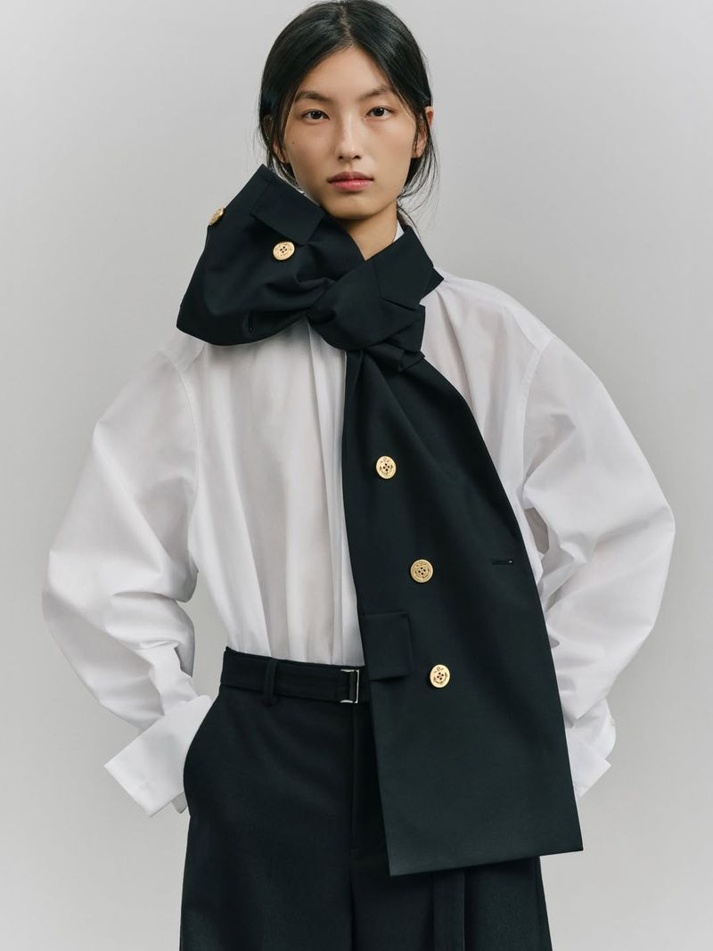 Asian woman wears a white shirt, black pants, and a black decorative neck piece with gold buttons in a studio shot.
