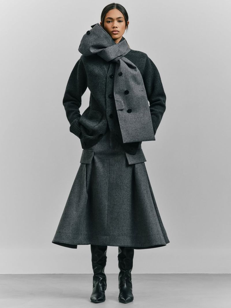 Fashionable woman in a stylish grey wool outfit, including a scarf and skirt, posing in a studio with a neutral backdrop, looking at camera.