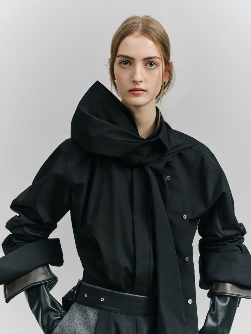 A woman poses in a stylish black shirt with an oversized collar, belted waist, and gray trousers against a pale gray backdrop.