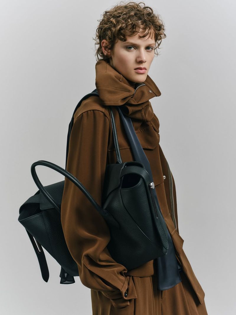 Fashionable woman with curly hair poses with a black leather tote bag in a photo studio wearing a brown coat and pants.