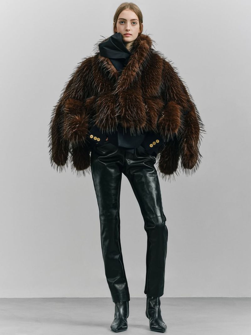 Full-length shot of a model wearing a shaggy brown fur coat with black leather pants and black boots against a neutral backdrop.