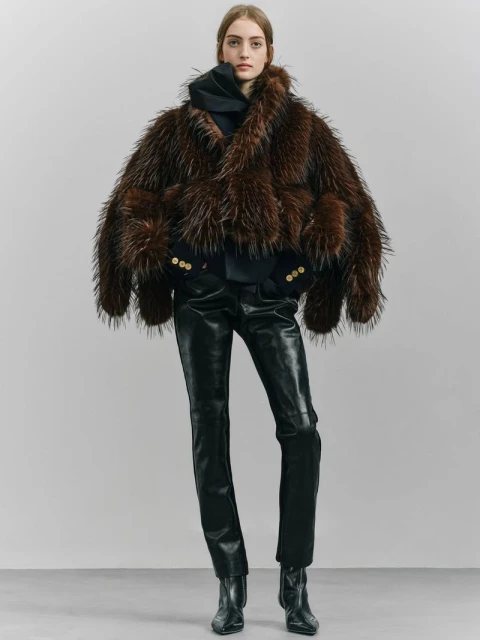 Full-length shot of a model wearing a shaggy brown fur coat with black leather pants and black boots against a neutral backdrop.