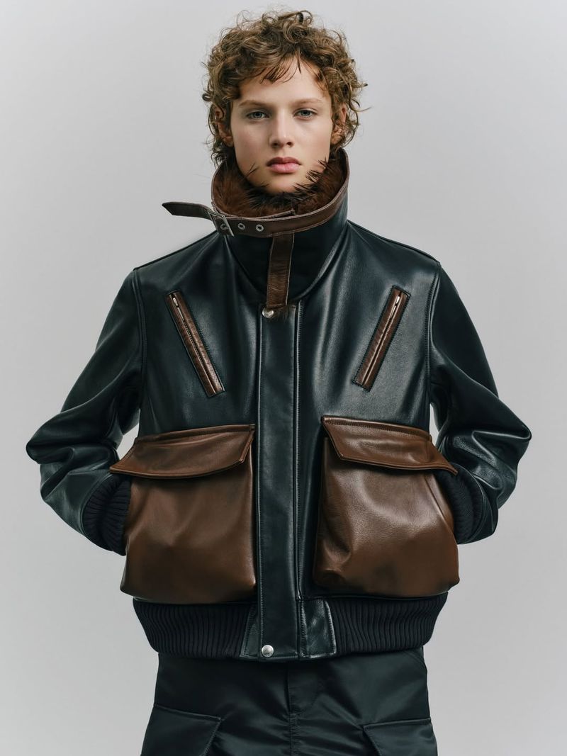 Person wearing a dark leather-look jacket with brown leather pockets and trim, against a light grey studio background. Fashion shot.