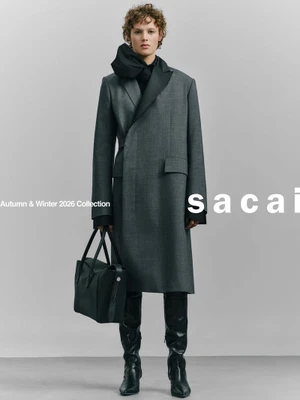 Model in grey coat and black boots holding a dark handbag against a grey seamless background for Sacai