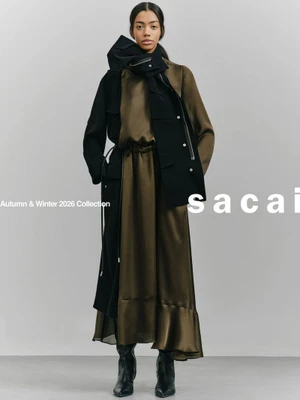 Model wearing an olive green satin dress and a black layered coat against a light grey studio background.
