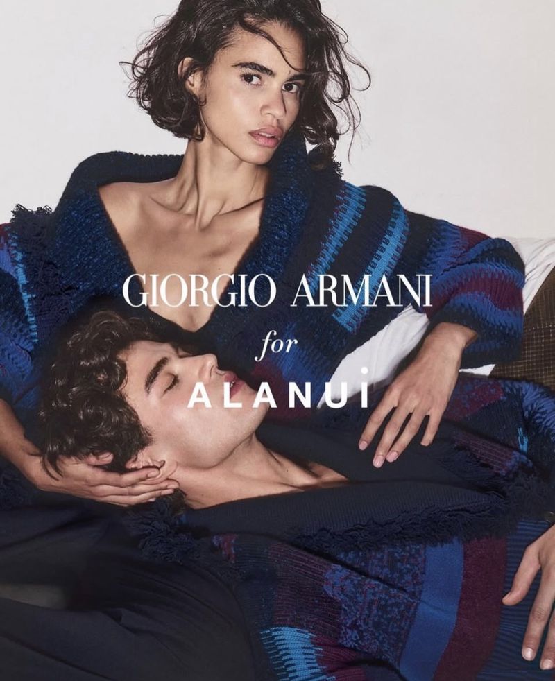 Two fashion models pose together in a studio setting, wearing a blue and brown knit sweater. The models have medium length dark hair.