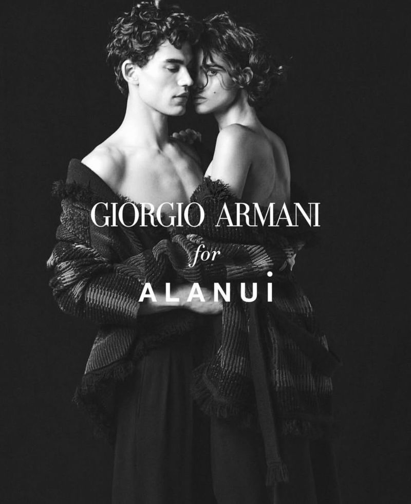 Black and white fashion image of two topless adult models embraced; wearing dark draped clothing, against a stark black studio backdrop.