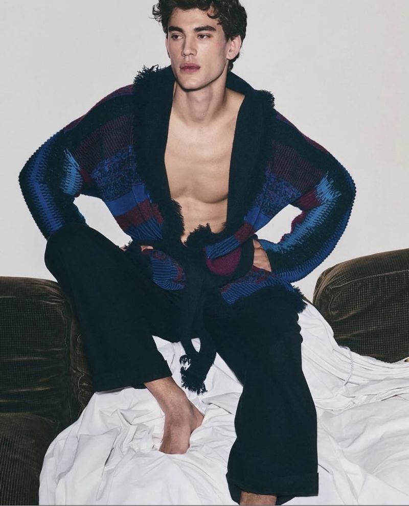 Male fashion model poses indoors in a patterned cardigan, dark pants, sitting on a couch covered with a white sheet in a fashion editorial.