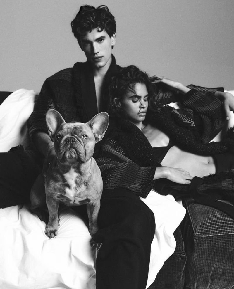 Black and white shot of an adult man and woman with a French bulldog on a bed in a photo studio, showcasing an editorial style.
