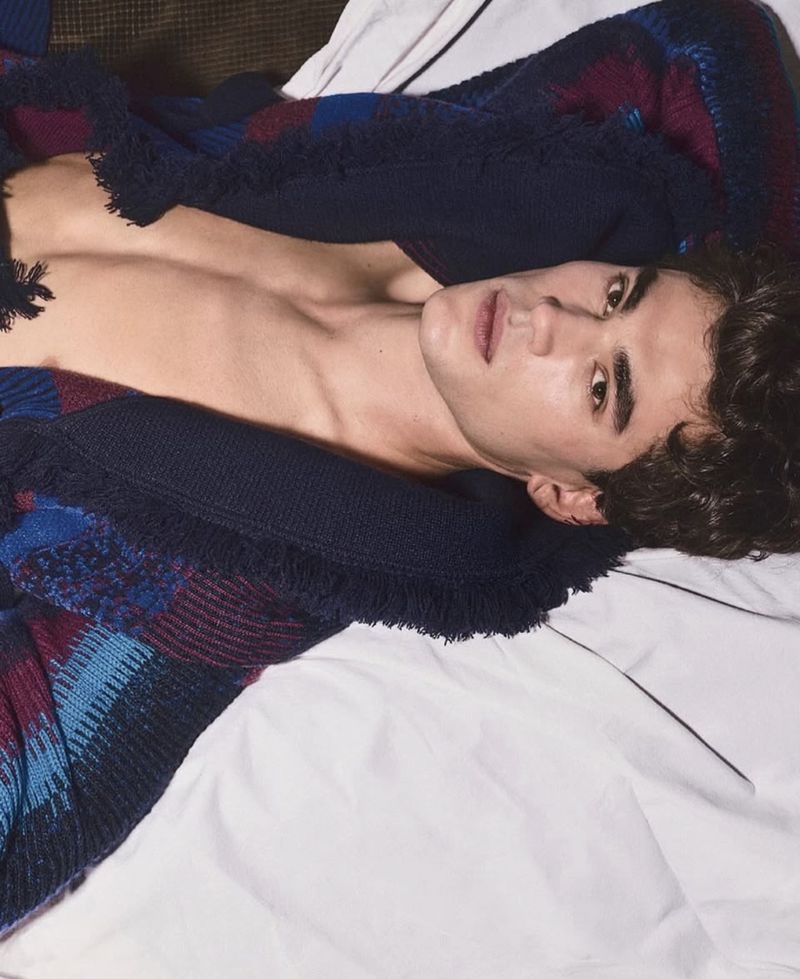 Young adult man lies on a white bed covered with a navy, blue, and burgundy patterned knit blanket. The lighting is soft and diffused.