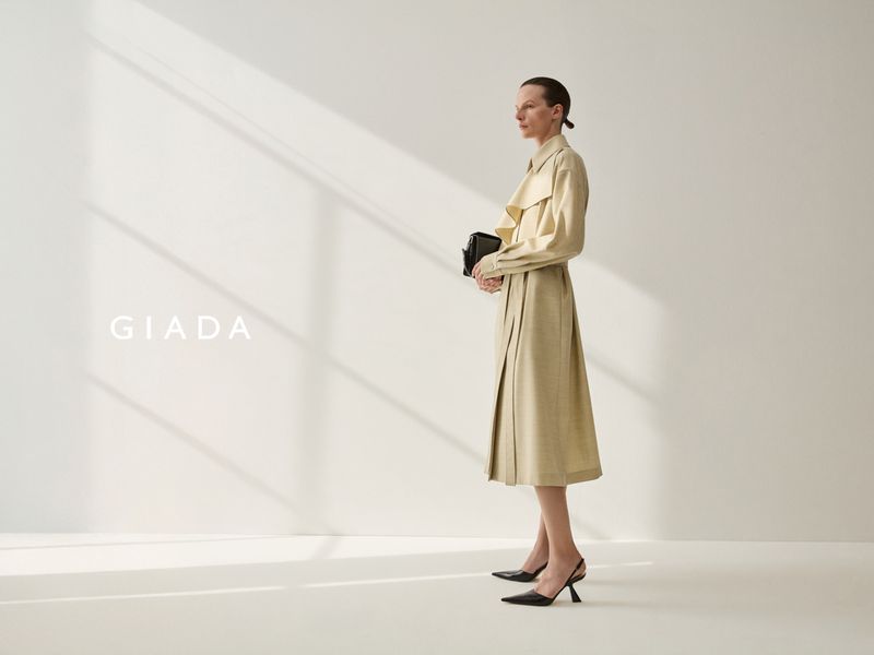 A woman in a tan trench coat holds a black clutch, standing in profile against a white wall with graphic shadows in a fashion shot.