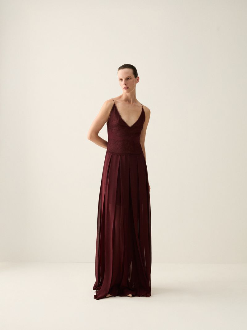 Elegant woman in a long, flowing burgundy dress with a V-neckline, posing in a bright, neutral-toned studio for a fashion editorial.