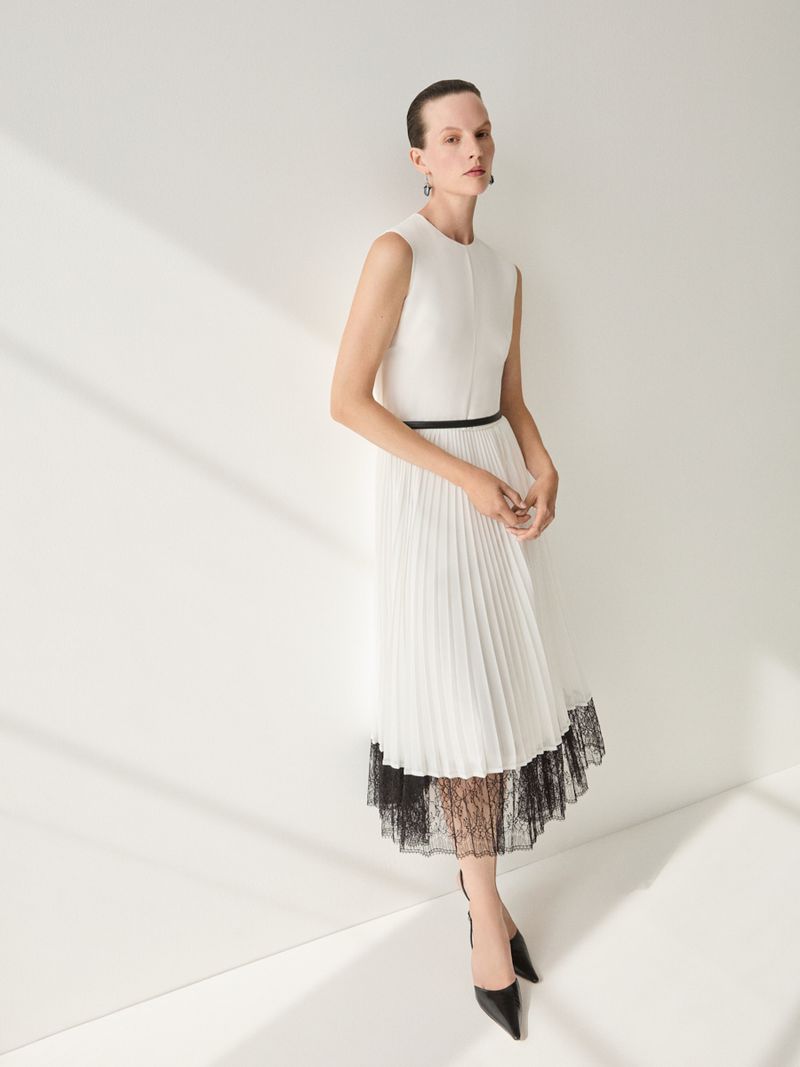 A woman in a white dress and black heels poses against a white wall with shadows, minimalist fashion and style, clean studio lighting.