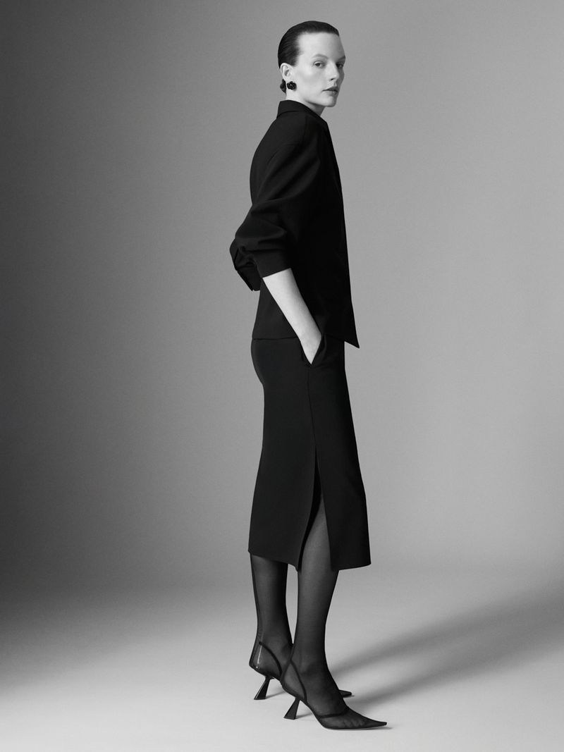 A woman in a black skirt suit and sheer black hose poses in a studio with a light grey background for a fashion editorial shot.