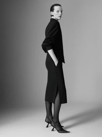 A woman in a black skirt suit and sheer black hose poses in a studio with a light grey background for a fashion editorial shot.