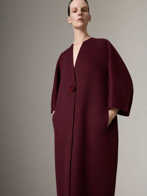 Fashion model in a mid-shot wearing a dark red coat with hands in her pockets against a solid light gray backdrop in a studio.