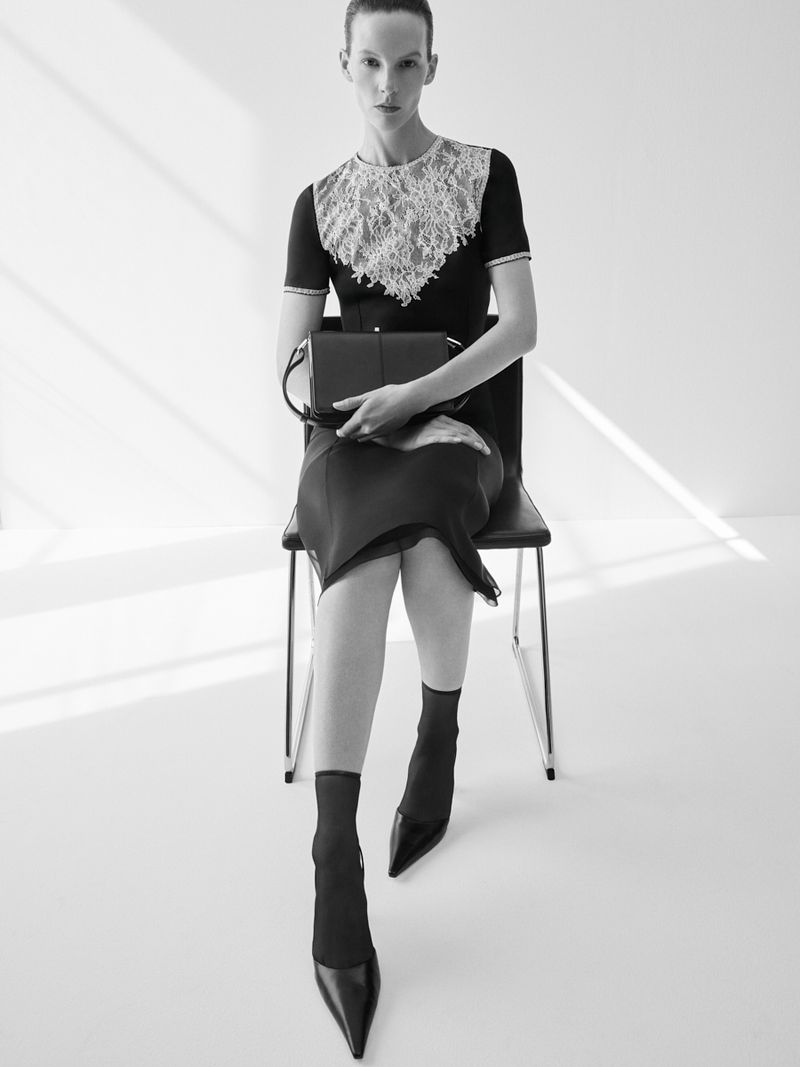Monochrome fashion shot: A slender model in a black dress with a lace collar, black hosiery, and heels, sitting on a chair in a bright studio.