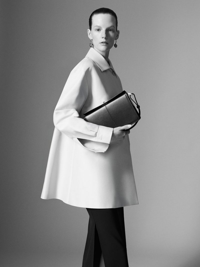 Monochrome fashion shot featuring an adult female model in a white jacket, black pants, and holding a black handbag in a studio setting.