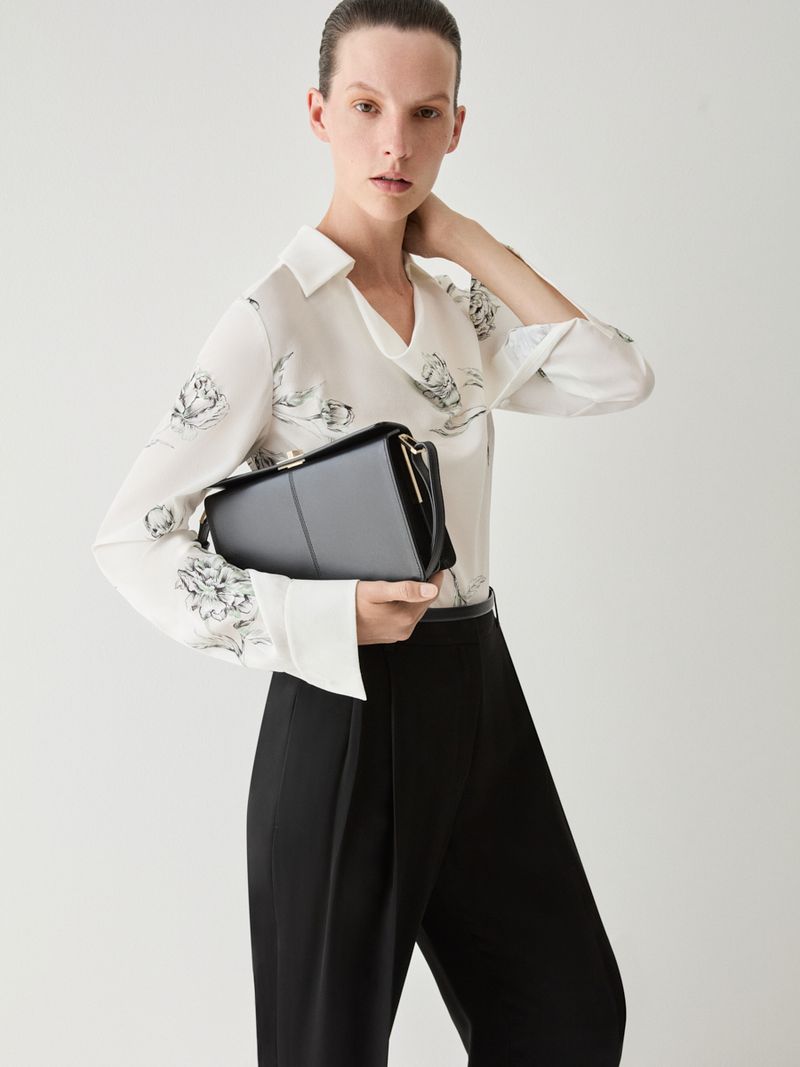 Elegant adult woman in long-sleeve floral shirt and black pants holding a glossy black purse in a studio setting with white background.