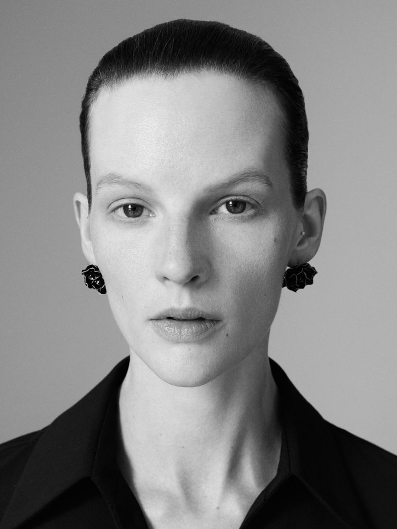 Close up portrait of a woman wearing black floral earrings and a dark collar against a neutral grey background