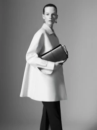 Model Sara Blomqvist holding a dark handbag wearing a white jacket against a grey studio seamless background.