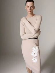 Model wearing a beige knit top and matching skirt with floral embroidery against a neutral grey studio background